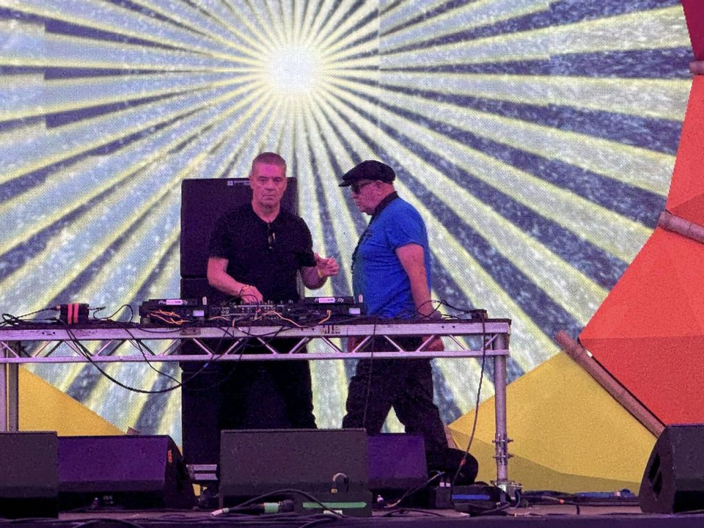 Gary Clail and Tony Wrafter on stage at Shambala 2024, projection of sun image behind them