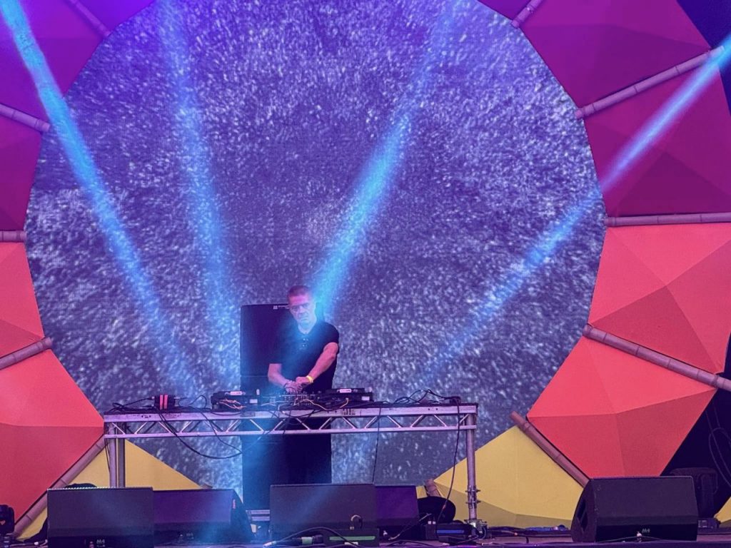 Gary Clail DJ'ing on stage at Shambala 2024, with trippy projection behind him
